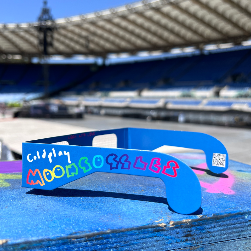 4: MoonGoggles: blue cardboard glasses. Coldplay is written in white. MoonGoggles is spelled out in green, blue, purple and pink. Each letter has a unique, playful hand-drawn style. On the inside of the glasses' arms, there’s a small pink message: LOOK WITH LOVE. The stage and stadium seats are in the background.