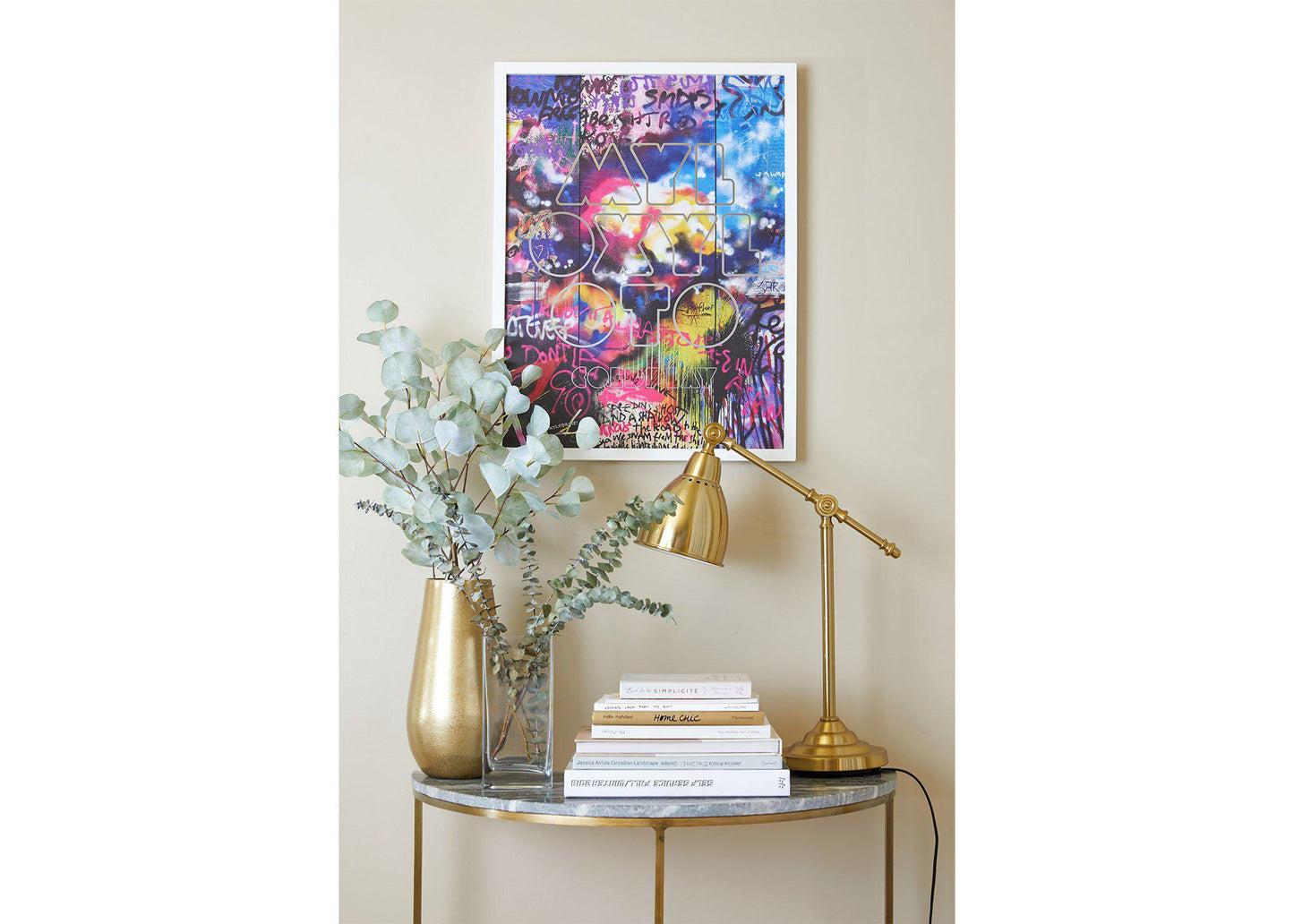 Mylo Xyloto Lithograph on a white frame, hung on a beige wall, above a small table shaped like a crescent moon, with a marble top and golden legs. The table has golden and clear vases with eucalyptus, books and a golden desk lamp. The poster's striking, expressive nature inspires rebellious energy and creativity