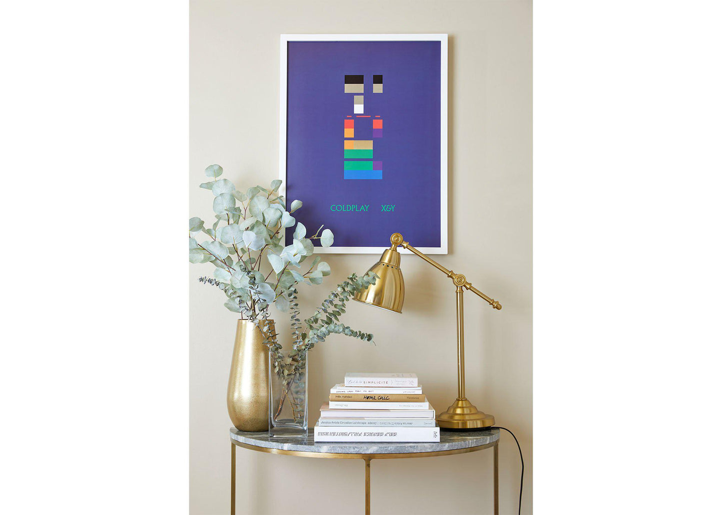 X&Y Lithograph on a white frame, hung on a beige wall, above a small table shaped like a crescent moon, with a marble top and golden legs. The table has golden and clear vases with eucalyptus, books and a golden desk lamp. The blocks’ red and orange harmonize with the golden, while The green and blue offer a refreshing contrast